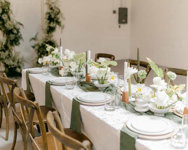 A long dining table is set with white dishes, candles, and floral arrangements, surrounded by wooden chairs in a simple venue with greenery in the background.