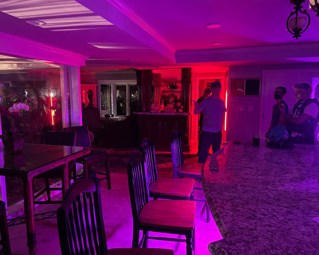 A dimly lit interior space with pink and purple lighting, highlighting a bar area and several chairs around a table.