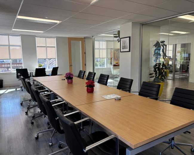 The image shows a modern conference room with a long wooden table surrounded by black chairs and large windows allowing natural light to enter.