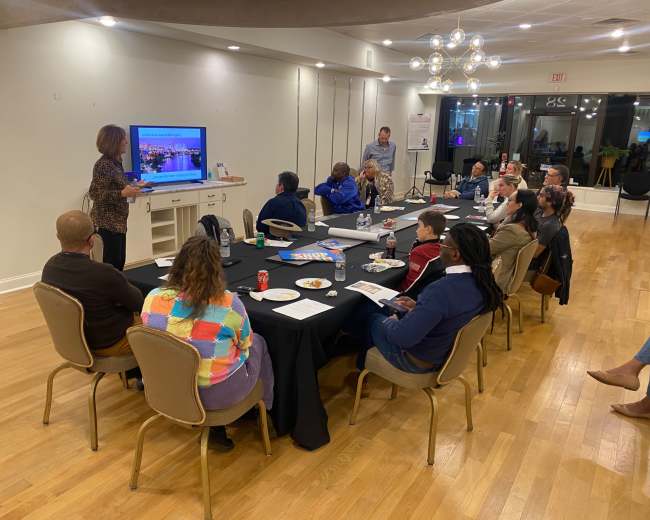 A group of people is seated around a long table in a conference room, attentively watching a presentation on a screen while snacks and drinks are on the table.