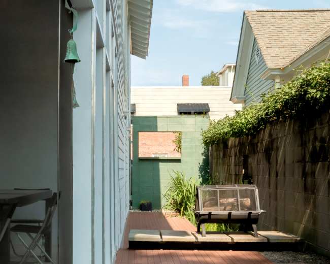 A narrow pathway leads between two buildings, with a garden area featuring a small table and a container, flanked by greenery and a decorative bell hanging on the left side.