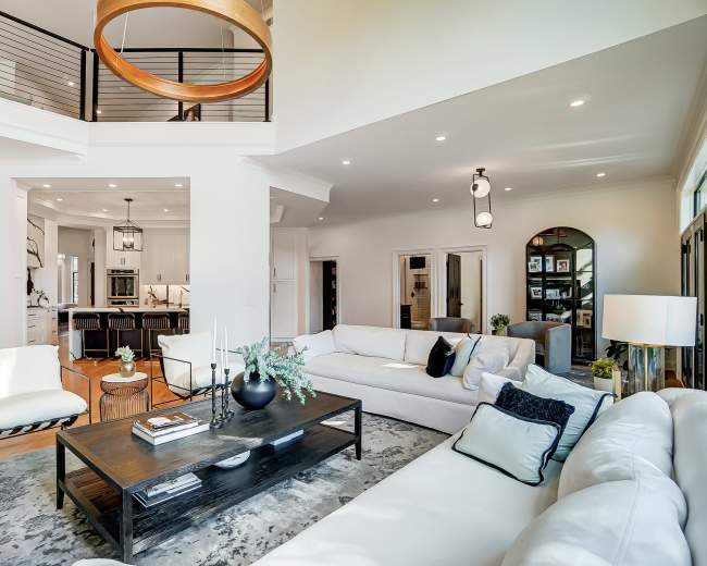 The image shows a modern living room with a white sectional sofa, a dark wooden coffee table, and large windows allowing natural light to fill the space.