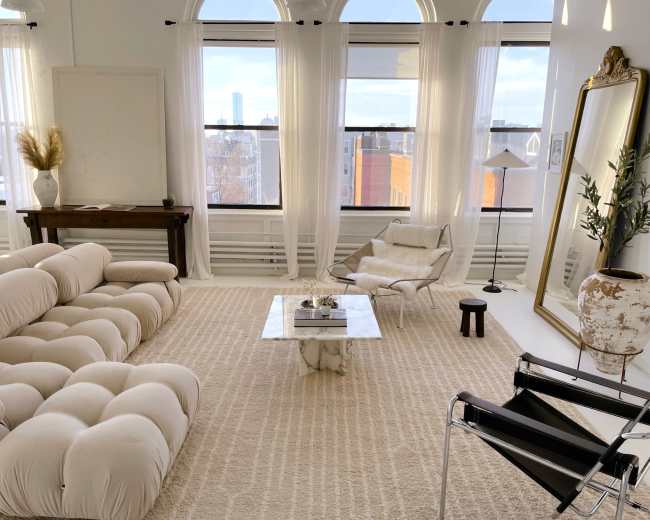 The image shows a modern living room featuring a beige sectional sofa, a glass coffee table, a large mirror, and large windows providing natural light.