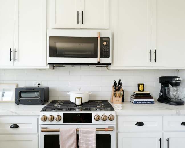 The image shows a modern kitchen with white cabinetry, a stainless steel microwave above a gas stove, and various kitchen appliances and tools arranged on the countertop.