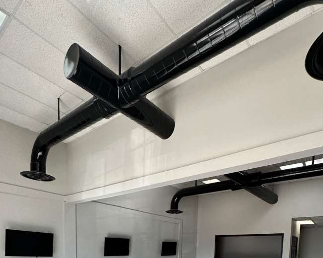 The image shows a spacious, undecorated room with high ceilings, large windows, and exposed black ductwork, featuring several flat-screen monitors mounted on the walls.