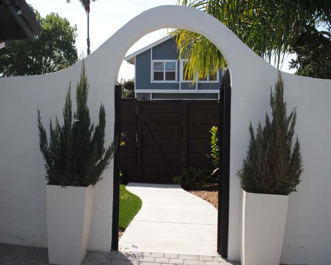 A white archway frames a path leading to a fenced garden area, with potted plants on either side.