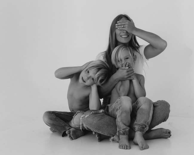 A woman sits on the floor with two children, playfully covering one child's eyes while the other covers their ears.