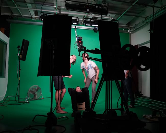 A group of people are positioned in a studio with a green screen, with one individual lying on a cart while others interact around them, and various filming equipment is visible in the foreground.