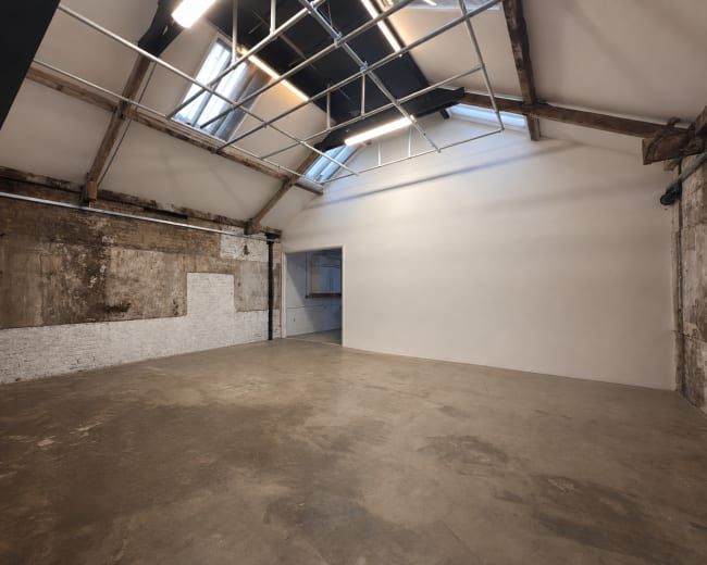 The image shows an open, empty studio space with exposed brick walls, large windows, and a high ceiling featuring wooden beams.
