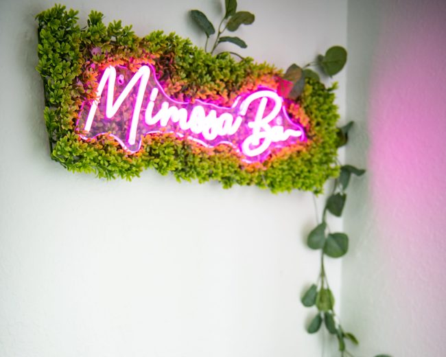 A wooden bar cart displays a chilled bottle of champagne, several empty glasses, and a pitcher under a neon sign that reads "Mimosa Bar."