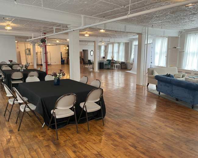 The image shows a spacious interior of a well-lit event space with tables covered in black cloth, chairs, and a seating area featuring a blue couch and large windows.
