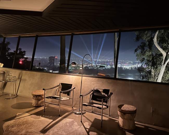 The image shows a modern interior with two chairs and a lamp, overlooking a city skyline illuminated by beams of light in the night sky.