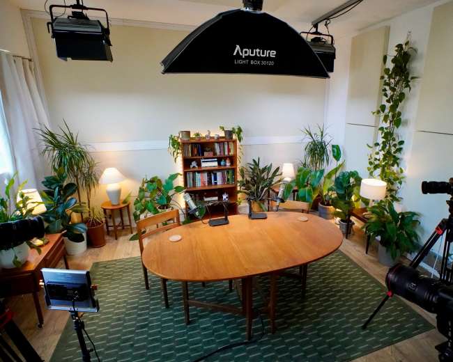 The image shows a well-lit studio setup for a discussion or interview, featuring a wooden table surrounded by chairs, plants, and cameras.