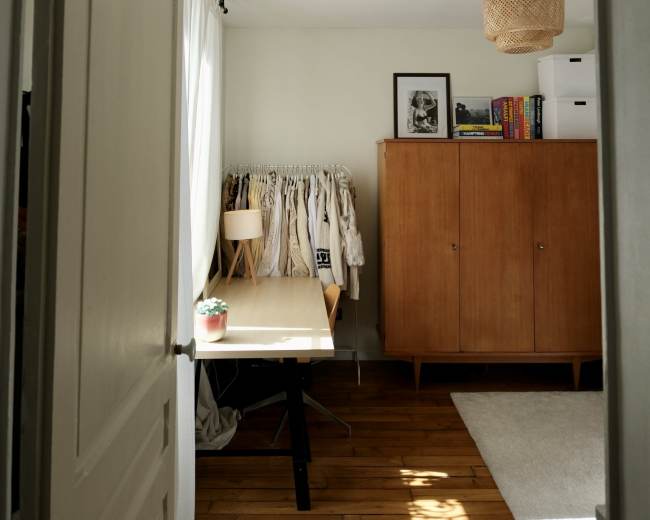 The image shows a well-lit room with a wooden floor, featuring a table, a wardrobe, and a clothing rack in the corner.