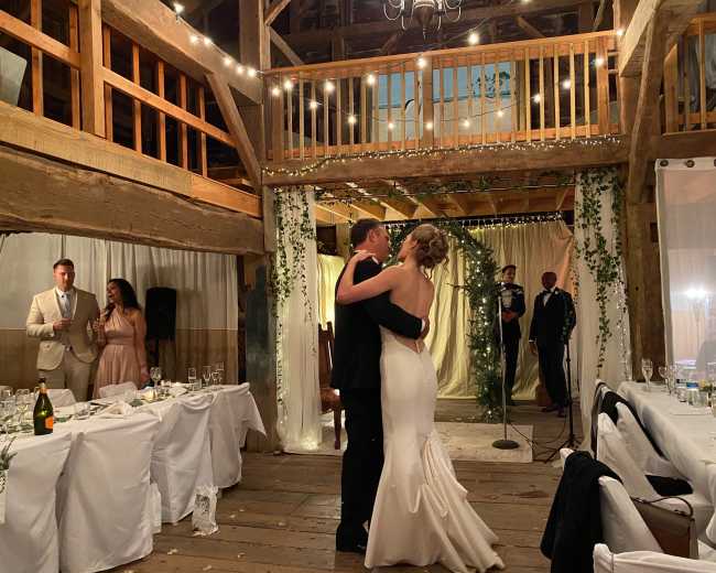 A couple dances in the center of a barn adorned with fairy lights and greenery, while guests observe from the side.