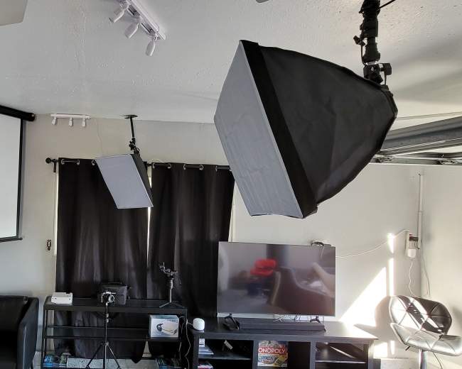 The image shows a modern living room setup featuring a large flat-screen TV, black curtains, a projector screen, and softbox lighting equipment.