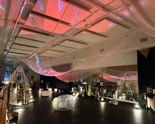 The image shows an art installation in a gallery featuring translucent fabrics suspended from the ceiling, interacting with soft lighting and various displayed sculptures.