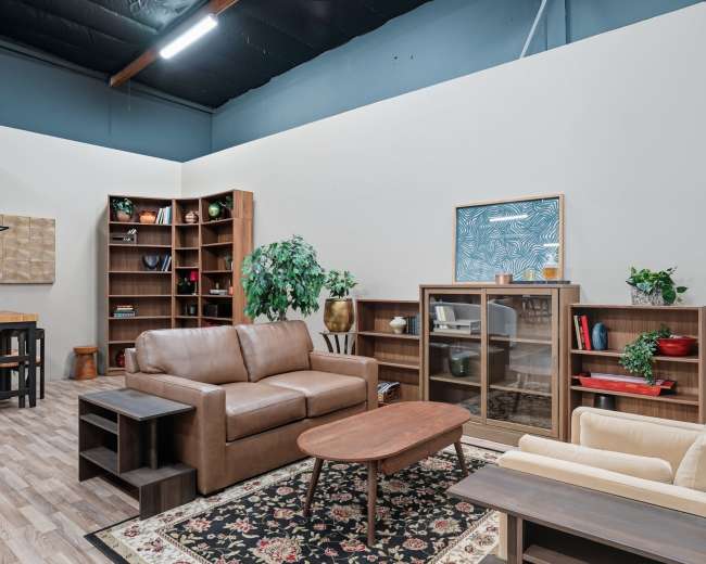 The image shows a cozy interior space featuring a brown leather sofa, a light-colored armchair, a wooden coffee table, and shelves filled with books and decorative items.
