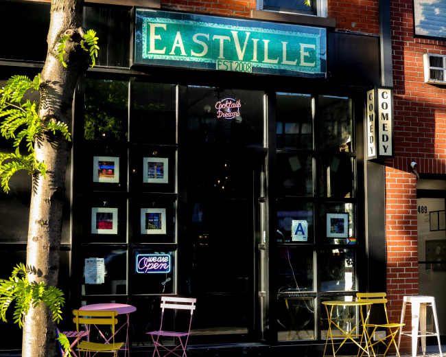 The image shows the exterior of a venue named "EastVille," featuring large glass windows with framed artwork, colorful chairs outside, and signage indicating it's open.