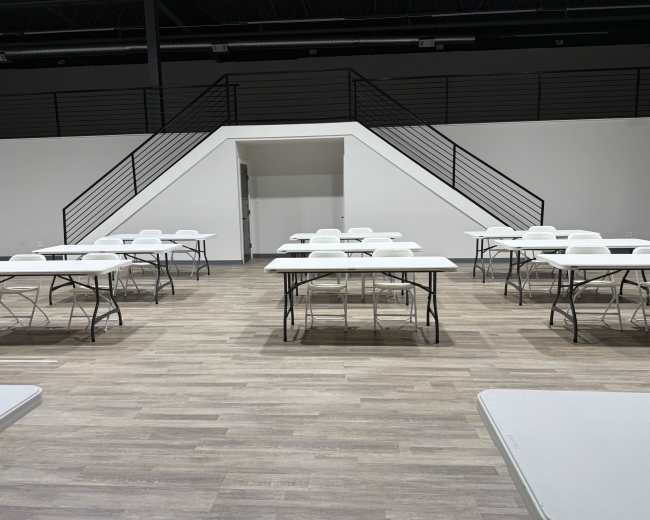 The image shows a spacious, empty room with several white tables set up in rows and a staircase in the background.