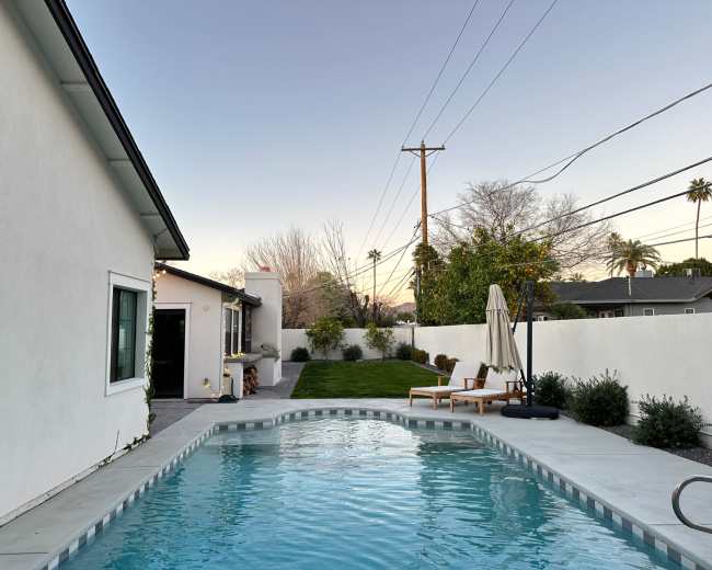 The image shows a modern backyard with a swimming pool, a lounge chair, and an umbrella, bordered by a white wall and surrounded by green landscaping.