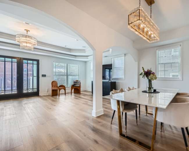 The image shows a bright dining area featuring a marble-topped table with upholstered chairs, adjacent to a living space with large windows and modern lighting fixtures.