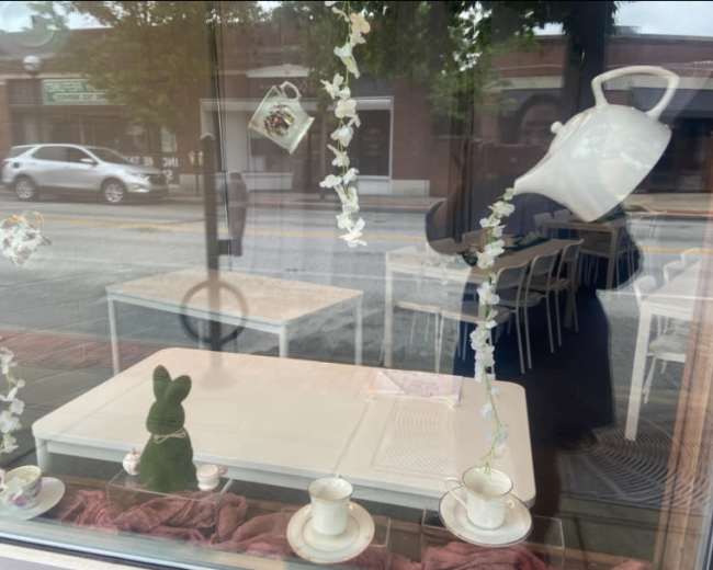 The image shows a storefront window display featuring a teapot, teacups, and decorative flowers, with a green teddy bear placed on a table.