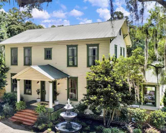 A two-story, light-colored house with green shutters and a front porch is surrounded by lush greenery and a fountain in the front yard.