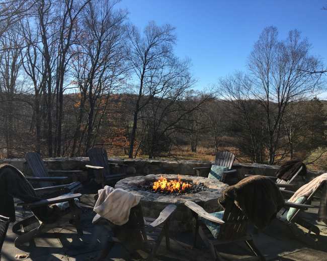 A stone fire pit surrounded by wooden chairs and blankets sits in a clearing with bare trees and a blue sky in the background.