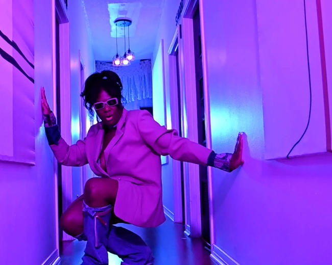 A person in a pink blazer and knee-high boots poses in a brightly lit hallway with purple hues.