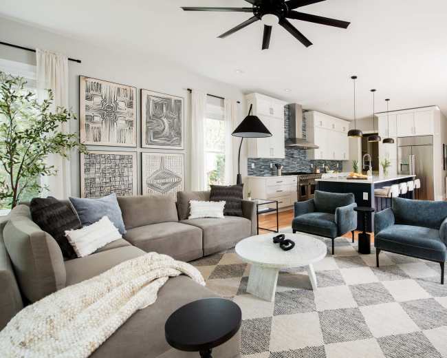 A modern living room features a large gray sectional sofa, a round coffee table, and patterned area rug, with artwork on the walls and a sleek kitchen visible in the background.