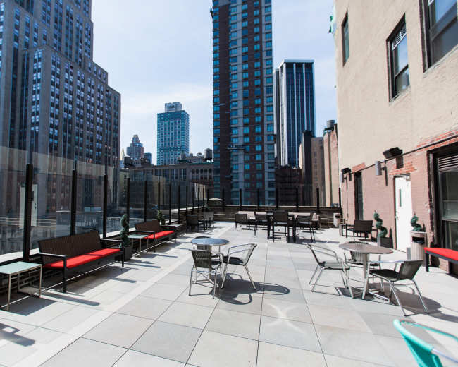 The image shows a spacious rooftop terrace with tables and chairs, surrounded by tall buildings in an urban setting.