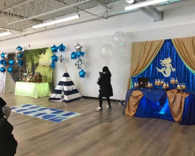 The scene features a decorated event space with blue and silver balloons, star-shaped decorations, and a themed table setup, including a blue tablecloth and a backdrop with an ocean-themed design.
