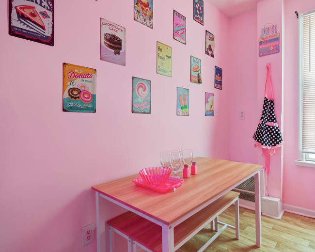 A small dining area features a light wood table with two benches and pink walls adorned with vintage food-themed signs.