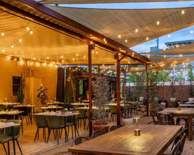 The image shows an outdoor dining area with string lights and shaded tables arranged around a central space.