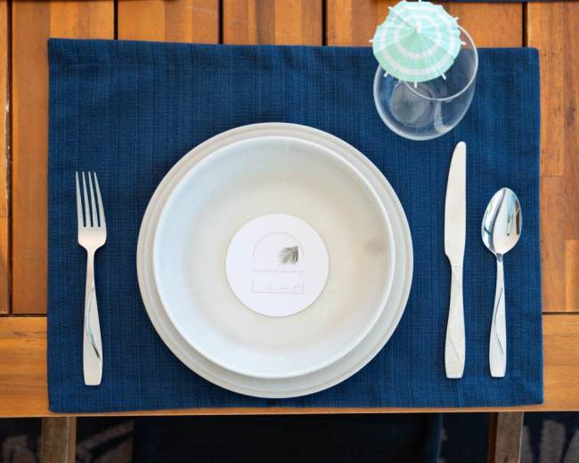 A setting featuring a blue placemat with a circular plate, a menu card labeled "Lead," a fork, knife, and a glass on a wooden table.