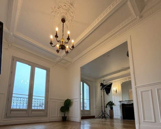 The image shows a well-lit, spacious room with wooden herringbone flooring, elegant molding on the ceiling, and large windows.