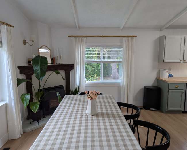A dining table with a checkered tablecloth is positioned in a bright kitchen, which features a large window and a green cabinet.