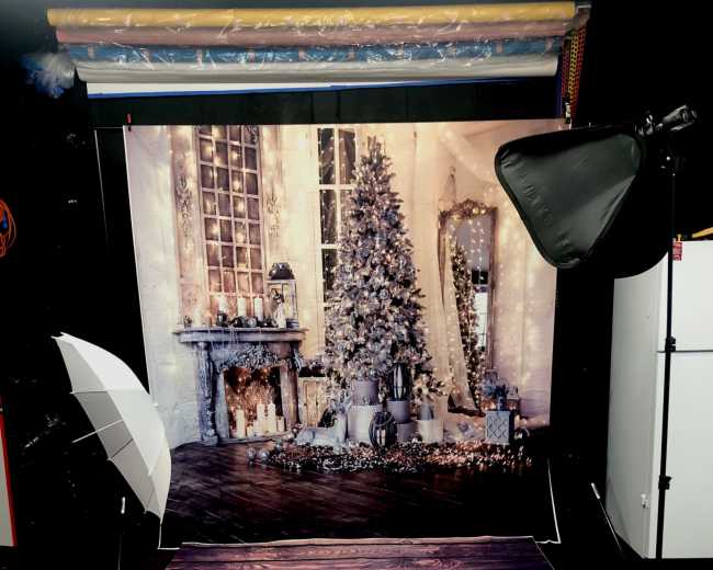 A photography setup featuring a decorated backdrop of a cozy room with a Christmas tree, flanked by two softbox lights on either side.
