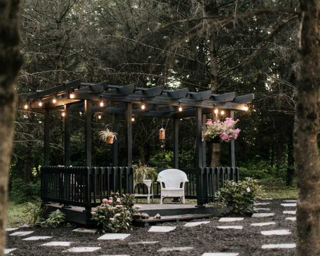 A black wooden gazebo with string lights and flower pots sits on a pathway surrounded by trees.