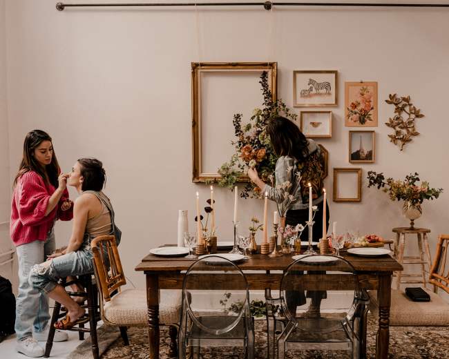 A dining table is set with candles and floral decorations, while one person applies makeup to another and another arranges flowers on a wall.