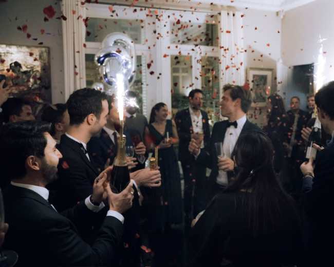 A group of elegantly dressed people celebrate at a party, holding champagne glasses and surrounded by confetti.