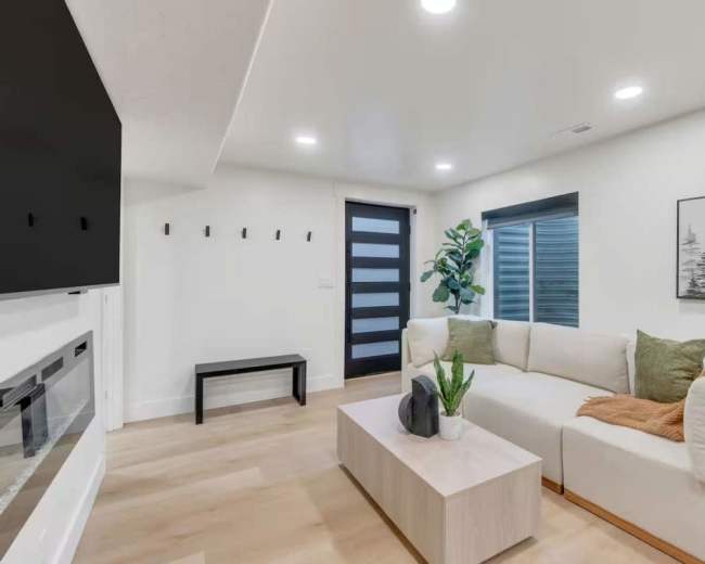 The image shows a modern living room featuring a sectional sofa, a coffee table, a plant, and a flat-screen television mounted on the wall.