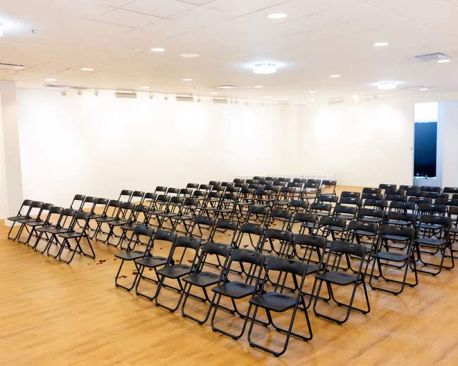 A large room features rows of black folding chairs arranged in a semi-circle facing a blank wall.