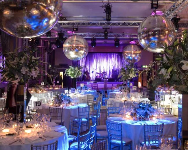 The image shows a decorated banquet hall with tables set for an event, featuring large disco balls overhead and a stage in the background.
