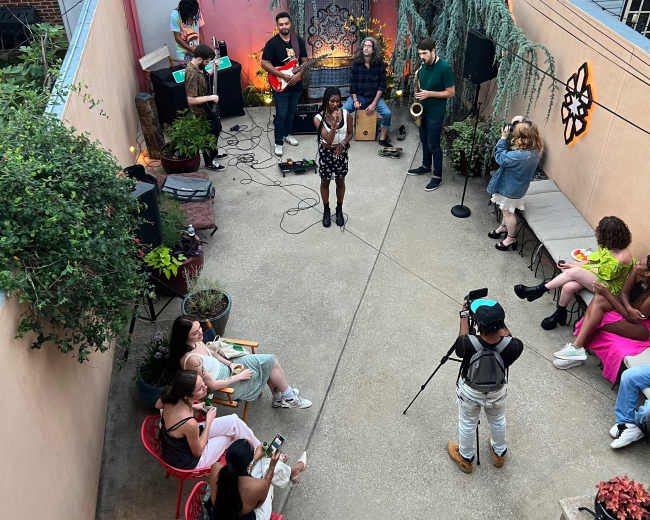A group of musicians performs on a small outdoor stage while an audience sits on chairs and benches in a courtyard adorned with plants.