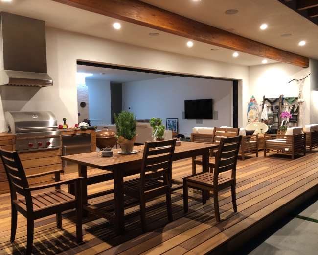 The image shows a modern outdoor dining area with a wooden table, chairs, a grill, and a seating arrangement under a covered patio.