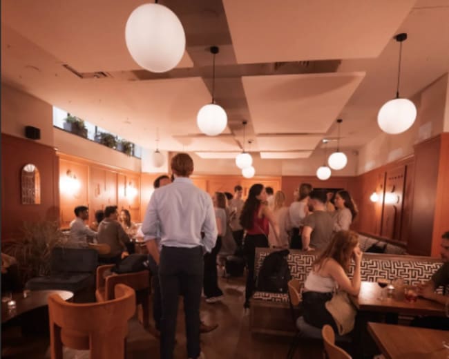 The image shows a bustling indoor restaurant with multiple groups of people socializing, warm lighting, and a mix of seating arrangements.