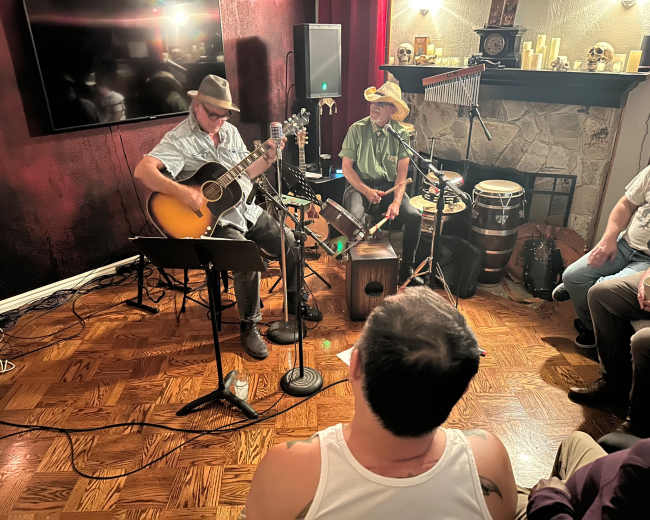 Two musicians perform in a cozy setting, one playing guitar while the other accompanies on percussion, with an audience seated in front.