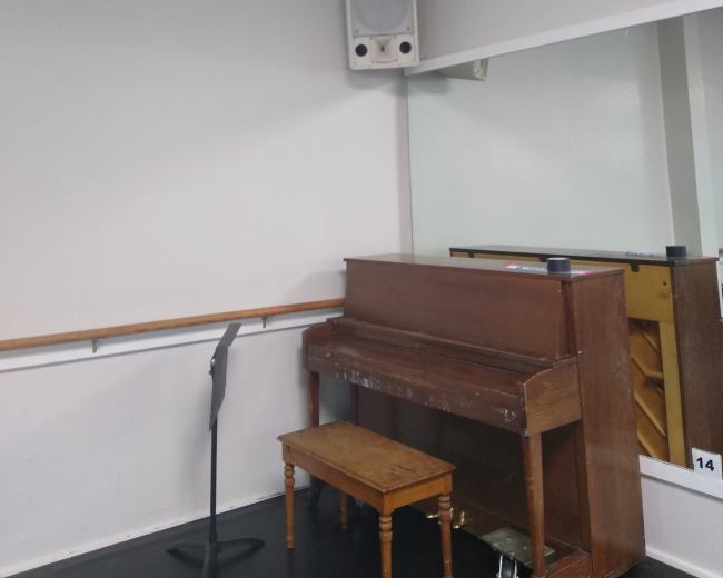 A wooden piano placed next to a small stool, with a wall-mounted speaker and a mirror on the opposite wall in a practice room.